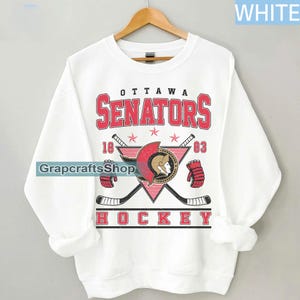 May include: White sweatshirt featuring the Ottawa Senators hockey team logo. The logo includes the team name in red, a stylized gladiator helmet, crossed hockey sticks, and the text "HOCKEY". The sweatshirt is hanging on a wooden hanger.