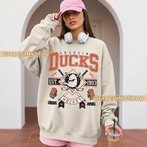 May include: Cream Anaheim Ducks sweatshirt featuring an orange distressed graphic. The design includes the team name, a hockey mask, crossed hockey sticks, and the text "EST. 1993" and "HOCKEY".