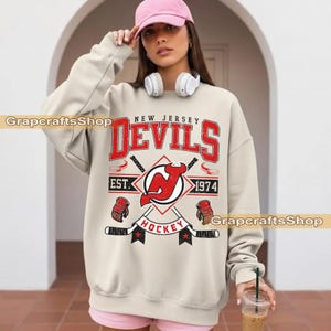 May include: Cream-colored sweatshirt featuring a red and black graphic design. The design includes the words "New Jersey Devils," "Hockey," and "Est. 1974," along with hockey-related imagery. The sweatshirt has long sleeves.