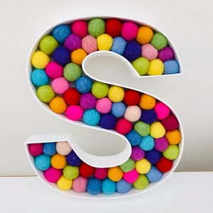 May include: A white letter S decorated with colourful felt balls. The letter is made of cardboard and has a three-dimensional shape.