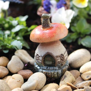 May include: A miniature mushroom-shaped house with a red roof and a chimney. The house is made of ceramic and has a detailed design. It is surrounded by small stones and green plants.