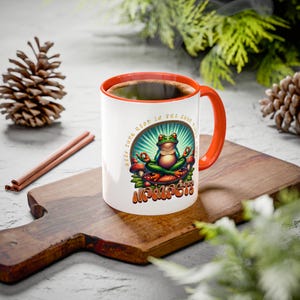 Puede incluir: Taza de cerámica blanca con asa e interior naranja, con un diseño de rana meditando. El diseño incluye la palabra "Namaste" y la frase "Keep Tone Good In The Good Times". La taza está llena de café y está sobre una tabla de cortar de madera.