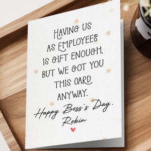 May include: A white greeting card with black text that reads: Having us as employees is gift enough, but we got you this card anyway. Happy Boss's Day. Robin. The card features small light orange stars and a tiny red heart at the bottom.