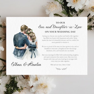 Puede incluir: Una tarjeta de boda rectangular con una ilustraci&oacute;n en acuarela de una pareja abraz&aacute;ndose. El texto dice: TO OUR Son and Daughter-in-Law ON YOUR WEDDING DAY, seguido de un mensaje, los nombres Ethan & Amelia y With all our love, Mom & Dad.