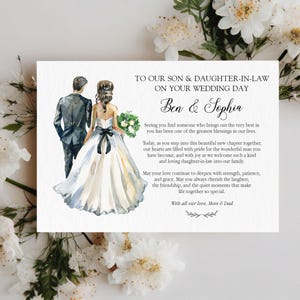 Puede incluir: Una tarjeta de boda rectangular con una ilustraci&oacute;n en acuarela de una pareja con ropa formal. El texto dice: TO OUR SON & DAUGHTER-IN-LAW ON YOUR WEDDING DAY, Ben & Sophia, seguido de un mensaje y firmado con With all our love, Mom & Dad.