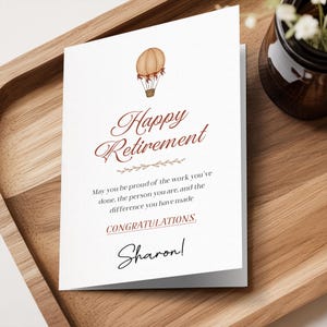 May include: A white greeting card featuring a brown hot air balloon illustration at the top. The text reads: Happy Retirement, May you be proud of the work you've done, the person you are, and the difference you have made, CONGRATULATIONS, Sharon!