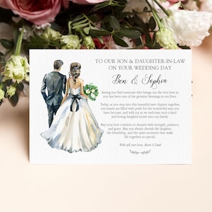 Puede incluir: Una tarjeta de boda rectangular con una ilustraci&oacute;n en acuarela de una pareja vista desde atr&aacute;s, el novio con traje oscuro y la novia con vestido blanco y faj&iacute;n negro. El texto dice: TO OUR SON & DAUGHTER-IN-LAW ON YOUR WEDDING DAY, Ben & Sophia, y un mensaje de amor de Mum & Dad.