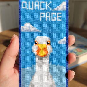 May include: A rectangular, blue cross-stitch design featuring a white goose with an orange beak and the words "QUACK PAGE" in white. The background is blue with white clouds. The design is framed with a dark blue border.
