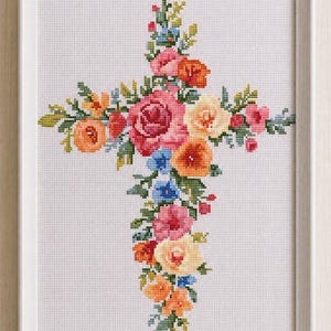 May include: A cross-stitch pattern featuring a floral cross design composed of various flowers in shades of pink, orange, yellow, and blue with green leaves, all contained within a simple light-colored rectangular frame on a white fabric background.
