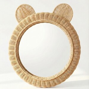 May include: A round, natural-colored rattan mirror with two rounded ears at the top, resembling a bear. The mirror has a woven texture and a circular shape, suitable for a nursery or children's room. The frame is made of woven rattan.