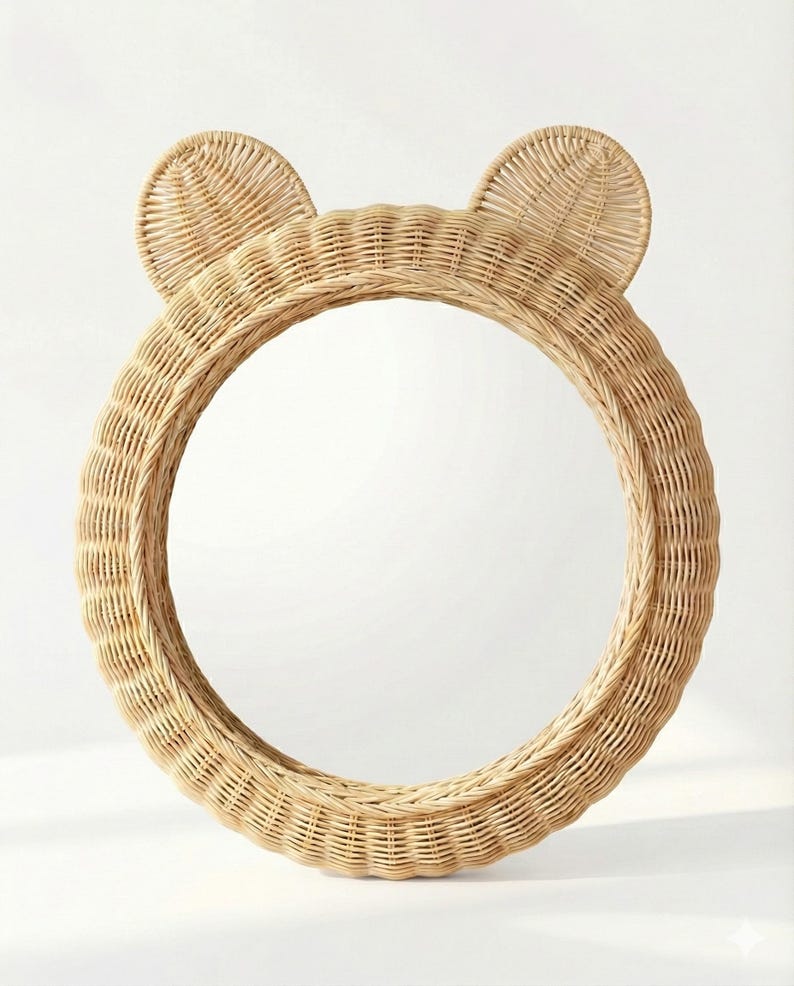 May include: A round, natural-colored rattan mirror with two rounded ears at the top, resembling a bear. The mirror has a woven texture and a circular shape, suitable for a nursery or children's room. The frame is made of woven rattan.