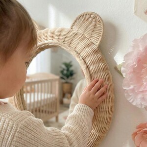 May include: A woven rattan mirror in the shape of a bear's head with ears. The mirror is round and hangs on a white wall. A child's hand touches the mirror. A large pink paper flower and a small bunny banner are also on the wall.