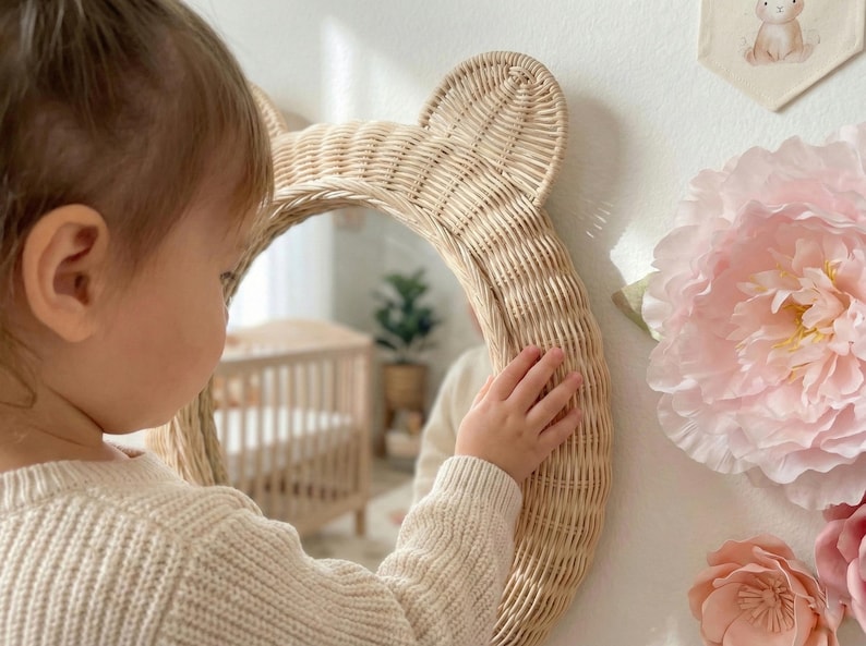 May include: A woven rattan mirror in the shape of a bear's head with ears. The mirror is round and hangs on a white wall. A child's hand touches the mirror. A large pink paper flower and a small bunny banner are also on the wall.
