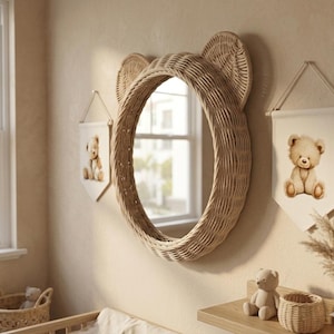 May include: A round wall mirror with a woven light brown rattan frame shaped like bear ears. Flanking the mirror are two white fabric wall banners, each featuring a printed illustration of a seated teddy bear. The mirror reflects a window.