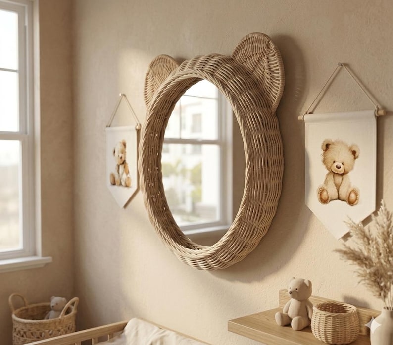 May include: A round wall mirror with a woven light brown rattan frame shaped like bear ears. Flanking the mirror are two white fabric wall banners, each featuring a printed illustration of a seated teddy bear. The mirror reflects a window.