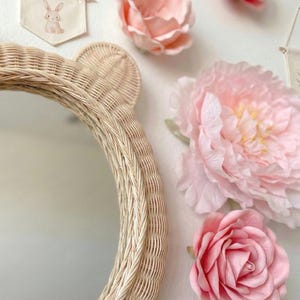 May include: A round, woven rattan mirror with bear ears. The mirror is surrounded by large, pink fabric flowers and small decorative banners with floral and bunny designs. The overall theme is a nursery or children's room.