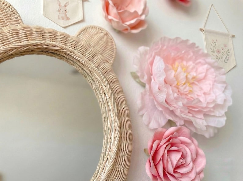 May include: A round, woven rattan mirror with bear ears. The mirror is surrounded by large, pink fabric flowers and small decorative banners with floral and bunny designs. The overall theme is a nursery or children's room.