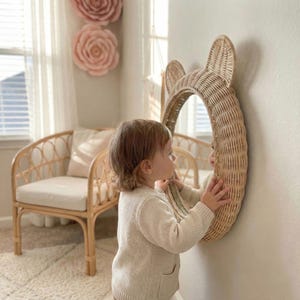 May include: A woven rattan mirror in the shape of a bear's head is mounted on a wall. The mirror is round and has two large, rounded ears at the top. The mirror is in a child's room, with a rattan chair and large paper flowers in the background.