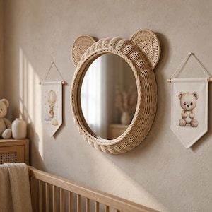May include: A round mirror framed in light brown woven rattan with two circular ears at the top. Flanking the mirror are two off-white fabric wall banners, one featuring a teddy bear in a hot air balloon and the other a seated teddy bear.