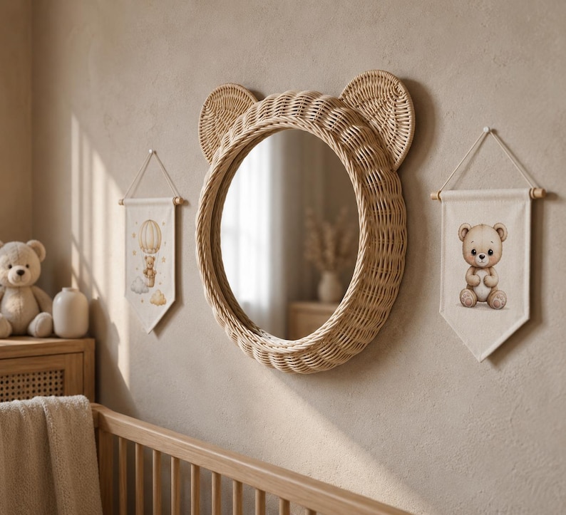 May include: A round mirror framed in light brown woven rattan with two circular ears at the top. Flanking the mirror are two off-white fabric wall banners, one featuring a teddy bear in a hot air balloon and the other a seated teddy bear.