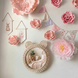 May include: A nursery wall decor arrangement featuring a round, woven mirror with bear ears, surrounded by large pink paper flowers, fabric banners with floral and bunny designs, and a pink bunting.