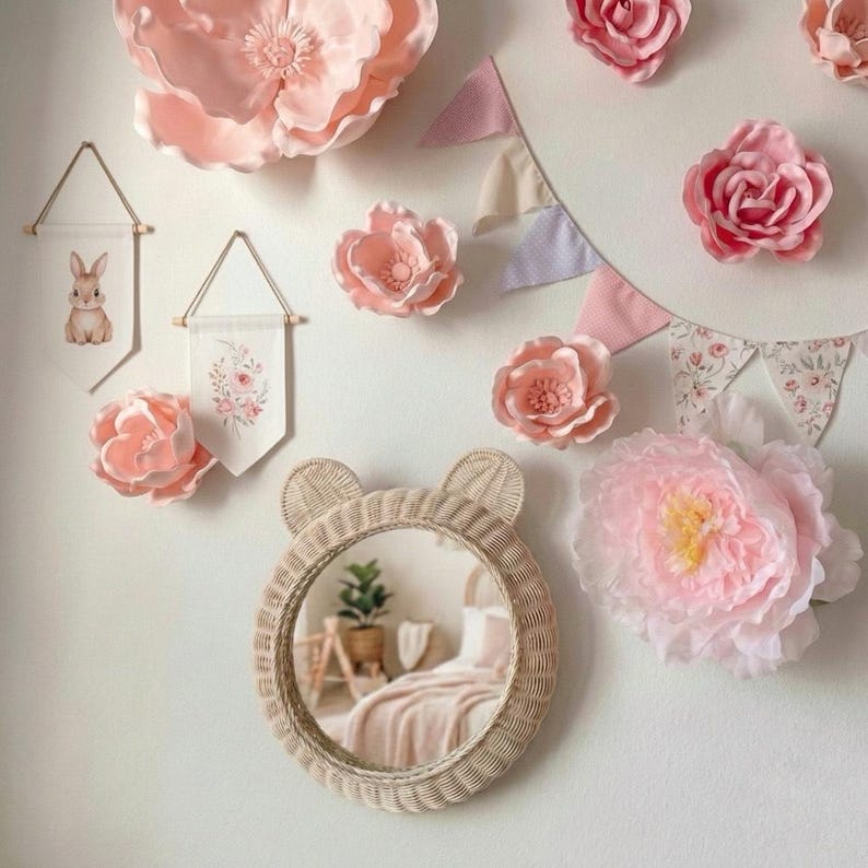 May include: A nursery wall decor arrangement featuring a round, woven mirror with bear ears, surrounded by large pink paper flowers, fabric banners with floral and bunny designs, and a pink bunting.