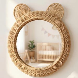 May include: A round, woven rattan mirror with two rounded ears at the top, resembling a bear. The mirror is a light tan color and is mounted on a white wall. The reflection shows a nursery with a crib and a plant.