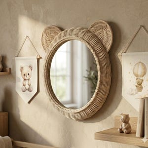 May include: A round wall mirror with a light brown woven rattan frame shaped like bear ears. The mirror is flanked by two fabric wall hangings, one featuring a teddy bear and the other a teddy bear in a hot air balloon, with a small bear figurine.