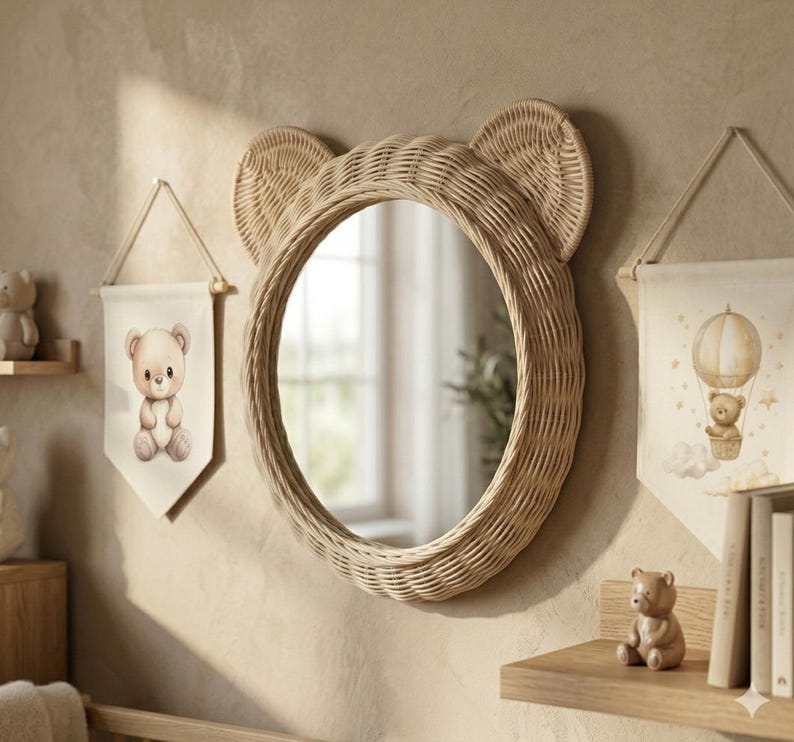May include: A round wall mirror with a light brown woven rattan frame shaped like bear ears. The mirror is flanked by two fabric wall hangings, one featuring a teddy bear and the other a teddy bear in a hot air balloon, with a small bear figurine.