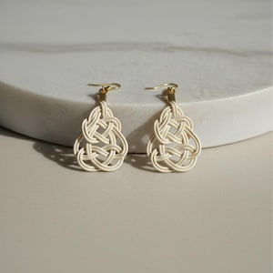 May include: A pair of off-white, woven earrings with a knotted design. The earrings have gold-colored hooks and a teardrop shape. The earrings are displayed on a white, curved surface.