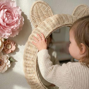 May include: A woven rattan mirror in the shape of a bear's head. The mirror is round and has two rounded ears at the top. The mirror is light beige in color and is hanging on a white wall. Decorative paper flowers are on the wall.