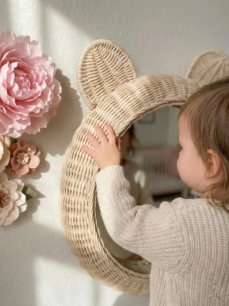 May include: A woven rattan mirror in the shape of a bear's head. The mirror is round and has two rounded ears at the top. The mirror is light beige in color and is hanging on a white wall. Decorative paper flowers are on the wall.