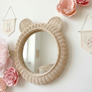May include: A round, woven rattan mirror with bear ears, hanging on a white wall. The mirror is surrounded by pink paper flowers and two small fabric banners with illustrations. The mirror's diameter is approximately 18 inches.