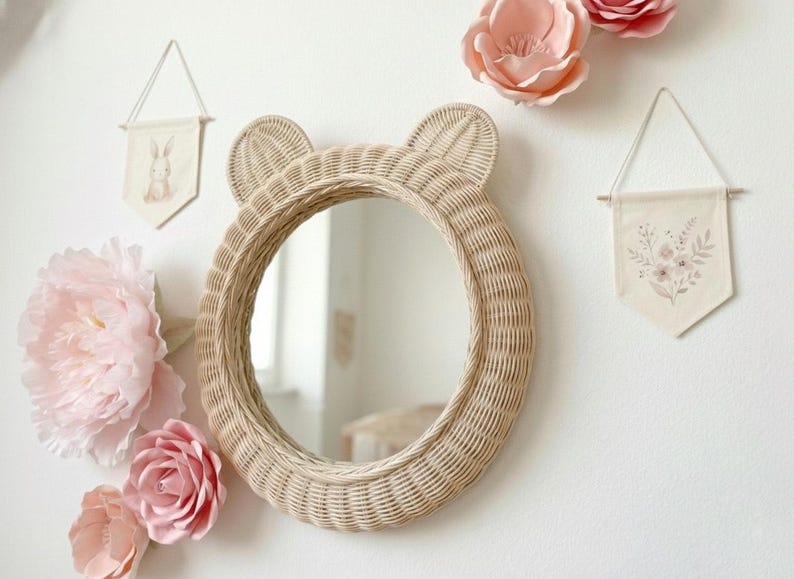 May include: A round, woven rattan mirror with bear ears, hanging on a white wall. The mirror is surrounded by pink paper flowers and two small fabric banners with illustrations. The mirror's diameter is approximately 18 inches.