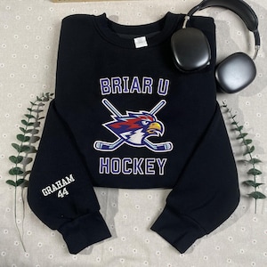May include: Black sweatshirt with the text "BRIAR U HOCKEY" and a logo of a bird with crossed hockey sticks. The sleeve has the name "GRAHAM" and the number "44" printed on it. A pair of black headphones is in the background.