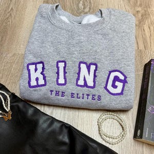 K&ouml;nnte beinhalten: Ein graues Sweatshirt mit dem Wort "KING" in wei&szlig;en Buchstaben mit lila Umrandung und dem Text "THE ELITES" in Lila. Eine schwarze Ledertasche, eine Perlenkette und ein Buch sind ebenfalls im Bild.