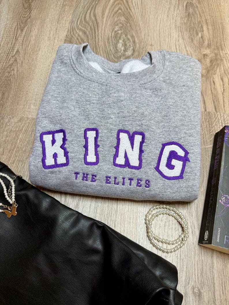 K&ouml;nnte beinhalten: Ein graues Sweatshirt mit dem Wort "KING" in wei&szlig;en Buchstaben mit lila Umrandung und dem Text "THE ELITES" in Lila. Eine schwarze Ledertasche, eine Perlenkette und ein Buch sind ebenfalls im Bild.