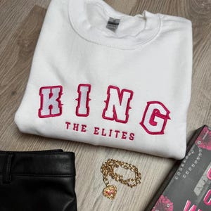 K&ouml;nnte beinhalten: Wei&szlig;es Sweatshirt mit dem Wort "KING" in Rosa gestickt, darunter "THE ELITES". Eine goldene Kette mit Herzanh&auml;nger, schwarze Hose und ein Buch sind ebenfalls im Bild.