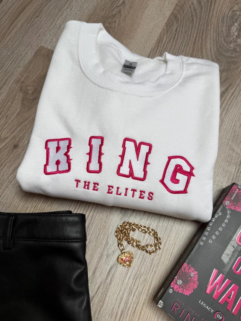 K&ouml;nnte beinhalten: Wei&szlig;es Sweatshirt mit dem Wort "KING" in Rosa gestickt, darunter "THE ELITES". Eine goldene Kette mit Herzanh&auml;nger, schwarze Hose und ein Buch sind ebenfalls im Bild.