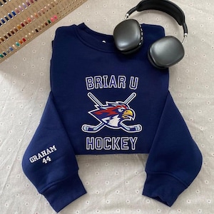 May include: A navy blue sweatshirt with the text "BRIAR U HOCKEY" and a logo of a hockey-playing eagle. The sleeve has "GRAHAM 44" embroidered in white. Black headphones rest on the sweatshirt.