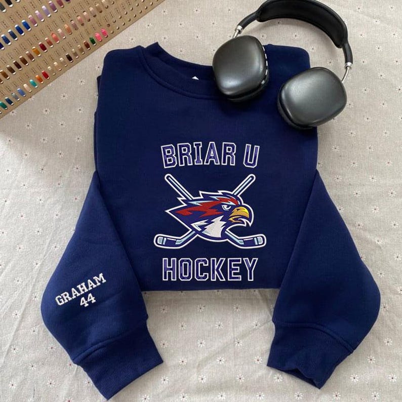 May include: A navy blue sweatshirt with the text "BRIAR U HOCKEY" and a logo of a hockey-playing eagle. The sleeve has "GRAHAM 44" embroidered in white. Black headphones rest on the sweatshirt.