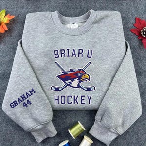 May include: A gray sweatshirt with the text "BRIAR U HOCKEY" and a graphic of a bird with crossed hockey sticks. The left sleeve has "GRAHAM 44" embroidered in blue. The sweatshirt is made of a soft material.