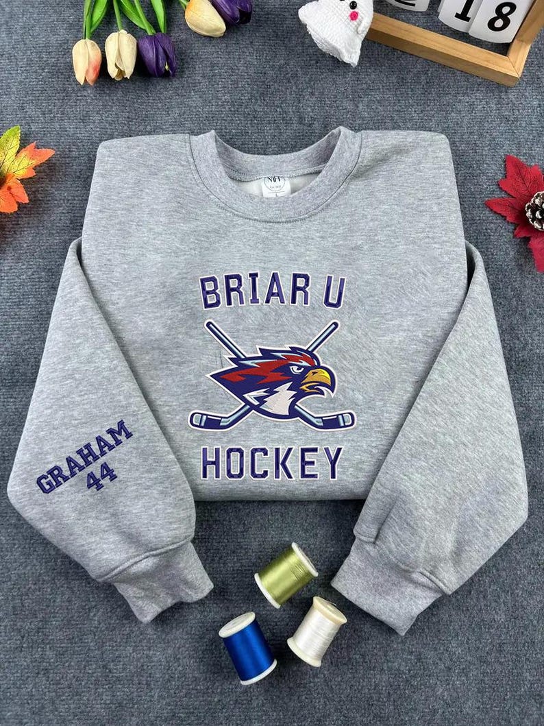 May include: A gray sweatshirt with the text "BRIAR U HOCKEY" and a graphic of a bird with crossed hockey sticks. The left sleeve has "GRAHAM 44" embroidered in blue. The sweatshirt is made of a soft material.