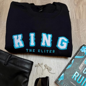 K&ouml;nnte beinhalten: Schwarzes Sweatshirt mit dem Wort "KING" in gro&szlig;en, hellblauen und wei&szlig;en Buchstaben. Unter dem Wort "KING" steht der Text "THE ELITES". Das Bild enth&auml;lt auch eine schwarze Sonnenbrille, ein Buch, eine schwarze Hose und schleifenf&ouml;rmige Ohrringe.