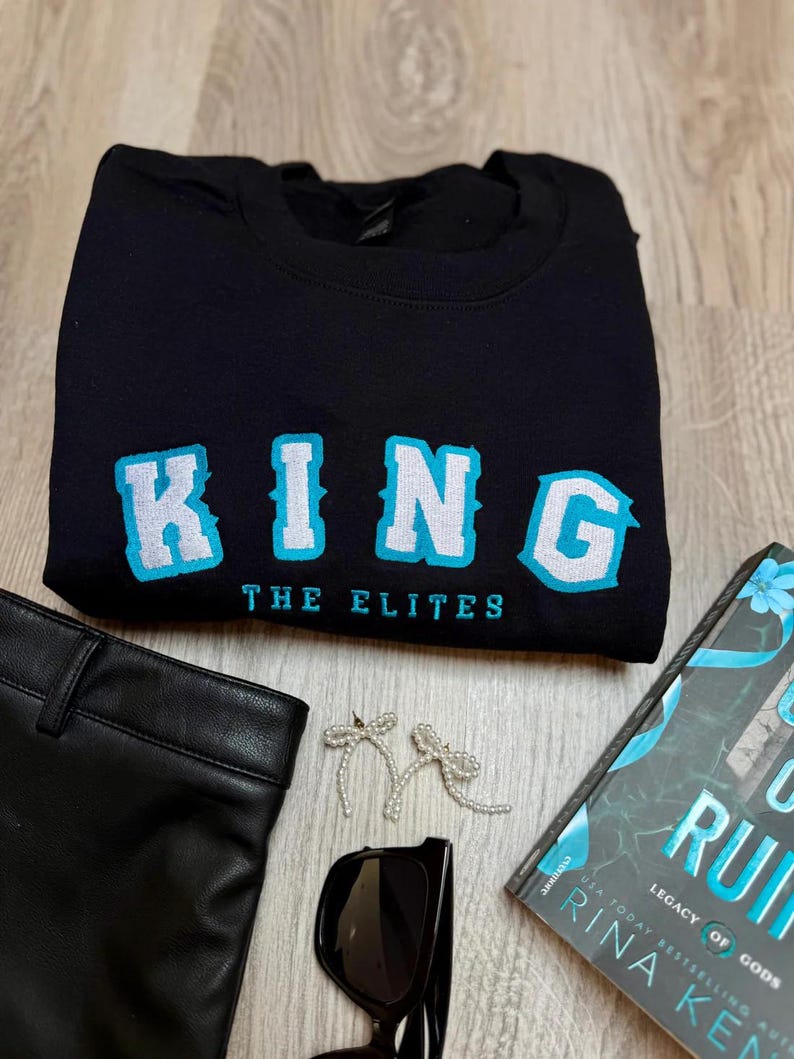 K&ouml;nnte beinhalten: Schwarzes Sweatshirt mit dem Wort "KING" in gro&szlig;en, hellblauen und wei&szlig;en Buchstaben. Unter dem Wort "KING" steht der Text "THE ELITES". Das Bild enth&auml;lt auch eine schwarze Sonnenbrille, ein Buch, eine schwarze Hose und schleifenf&ouml;rmige Ohrringe.