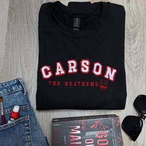 K&ouml;nnte beinhalten: Schwarzes Sweatshirt mit dem Namen "CARSON" in wei&szlig;en und roten Buchstaben. Darunter steht "THE HEATHENS" in Rot. Eine Sonnenbrille, ein Buch mit dem Titel "GOD OF MALICE" und eine Jeans sind ebenfalls im Bild.