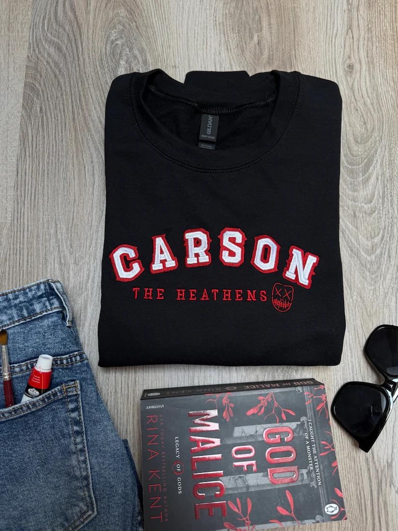 K&ouml;nnte beinhalten: Schwarzes Sweatshirt mit dem Namen "CARSON" in wei&szlig;en und roten Buchstaben. Darunter steht "THE HEATHENS" in Rot. Eine Sonnenbrille, ein Buch mit dem Titel "GOD OF MALICE" und eine Jeans sind ebenfalls im Bild.