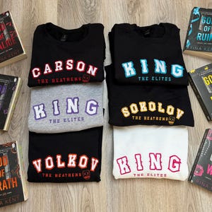K&ouml;nnte beinhalten: Eine Sammlung von Sweatshirts in Schwarz, Grau und Wei&szlig;, jedes mit fettem Text. Die Sweatshirts zeigen Namen wie "CARSON", "KING", "SOKOLOV" und "VOLKOV" sowie S&auml;tze wie "THE HEATHENS" und "THE ELITES".