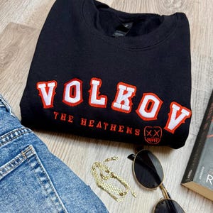 K&ouml;nnte beinhalten: Schwarzes Sweatshirt mit dem Wort "VOLKOV" in wei&szlig;er und roter Schrift. Unter dem Wort "VOLKOV" steht der Text "THE HEATHENS" und eine Grafik eines Gesichts mit einem X &uuml;ber den Augen. Das Sweatshirt wird mit Jeans, Sonnenbrille, einer goldenen Kette und einem Buch kombiniert.