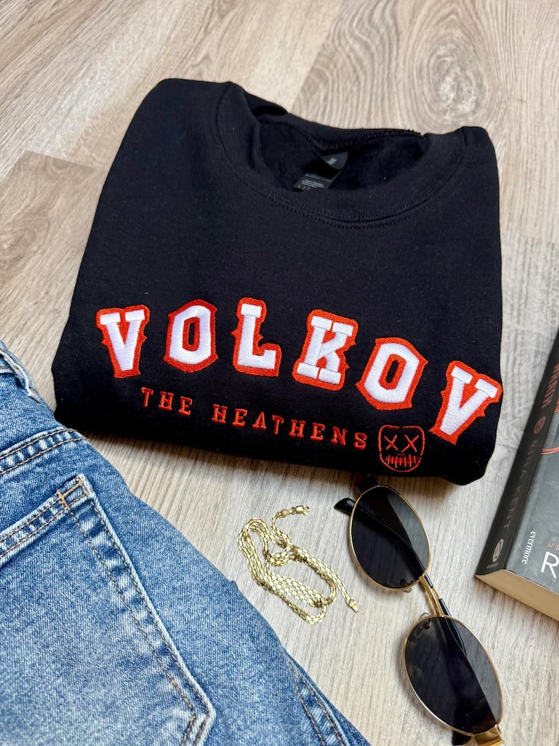 K&ouml;nnte beinhalten: Schwarzes Sweatshirt mit dem Wort "VOLKOV" in wei&szlig;er und roter Schrift. Unter dem Wort "VOLKOV" steht der Text "THE HEATHENS" und eine Grafik eines Gesichts mit einem X &uuml;ber den Augen. Das Sweatshirt wird mit Jeans, Sonnenbrille, einer goldenen Kette und einem Buch kombiniert.